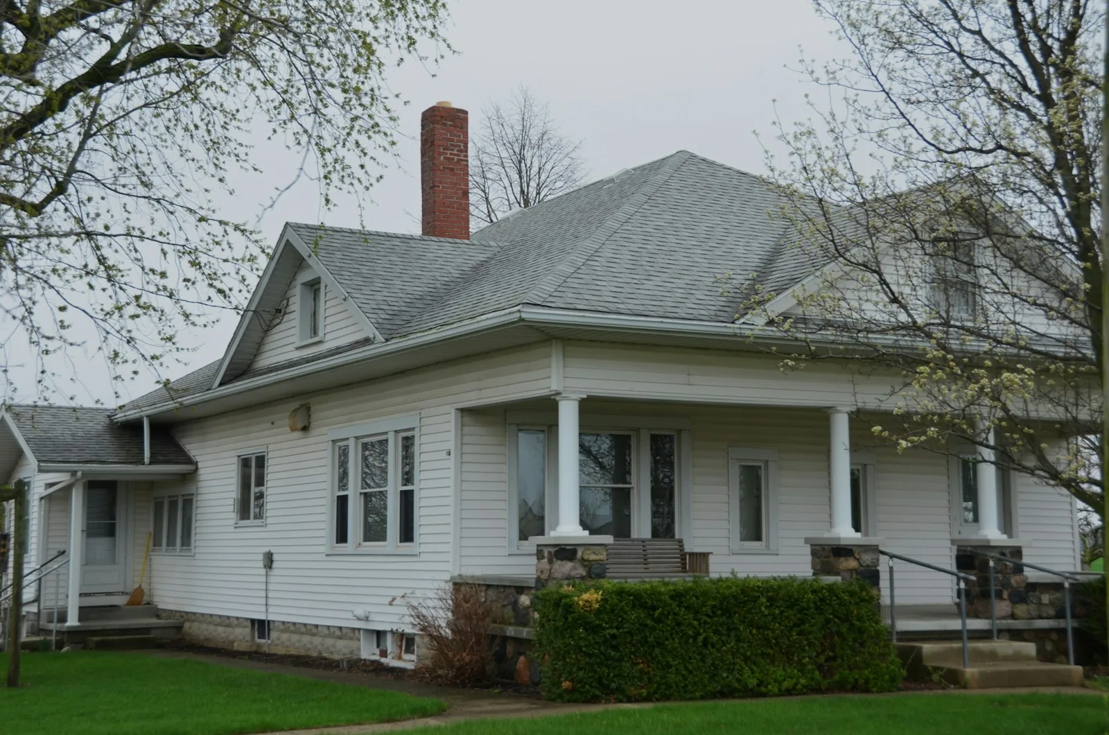 White hip-roofed bungalow with a brick chimney and covered porch under spring trees
